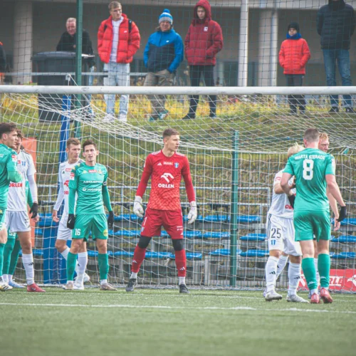 Na zdjęciu widać zawodników drużyny piłkarskiej stojących w pobliżu bramki, z bramkarzem w czerwonym stroju, podczas meczu na zielonym boisku, a w tle obserwujących mecz kibiców.