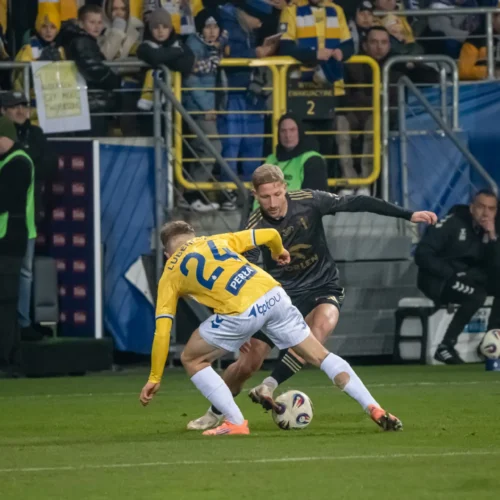 Na zdjęciu dwóch piłkarzy rywalizuje o piłkę podczas meczu, a w tle widać zebranych kibiców.