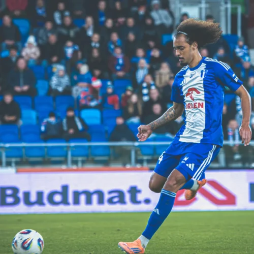 Na zdjęciu widać zawodnika w niebiesko-białym stroju piłkarskim, przygotowującego się do kopnięcia piłki na boisku, z widownią w tle.