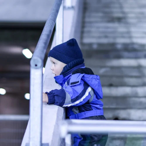 Na zdjęciu mały chłopiec w niebieskim stroju i czapce obserwuje coś zza balustrady w przestrzeni wewnętrznej.