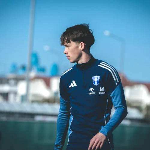Na zdjęciu widać młodego mężczyznę w niebieskim dresie sportowym, stojącego na boisku piłkarskim w słoneczny dzień.