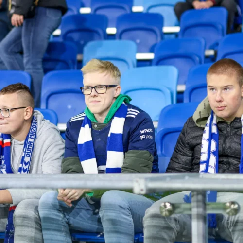 Na zdjęciu trzej młodzi mężczyźni w szalikach klubowych siedzą na trybunach stadionu, wpatrując się w boisko.