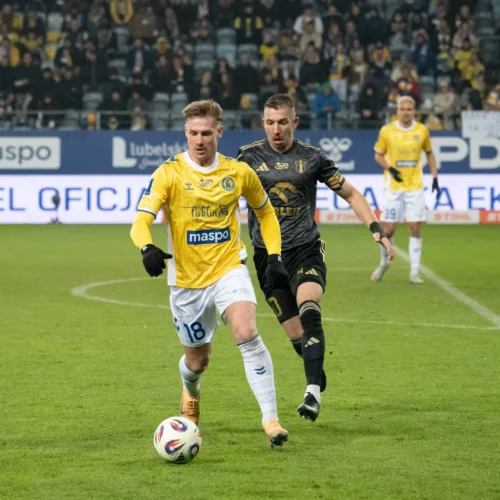 Na zdjęciu dwóch piłkarzy rywalizuje o piłkę na boisku, gdzie jeden z graczy w żółtej koszulce biegnie z piłką, a drugi w czarnej koszulce stara się go dogonić.