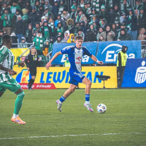 Na zdjęciu widać dwóch piłkarzy zmierzających do piłki na boisku podczas meczu w deszczowej aurze, z tłumem kibiców w tle.