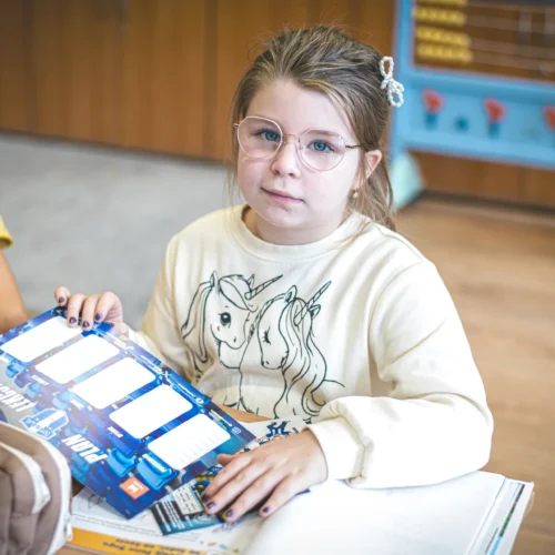 Na zdjęciu widać dziewczynkę w okularach, trzymającą planszę do gry, siedzącą w klasie obok chłopca.