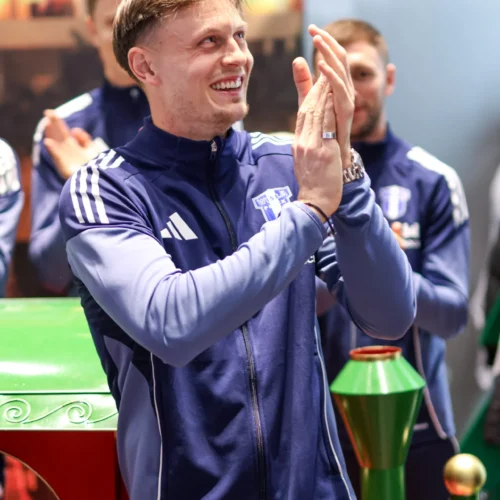 Na zdjęciu widać uśmiechniętego mężczyznę w sportowej odzieży, który klaskając, wyraża radość i entuzjazm na tle innych ludzi.