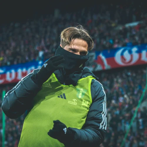 Na zdjęciu widać piłkarza w zielonej kamizelce na tle rozgrzanej atmosfery stadionu, który zamaskowany, wydaje gest w kierunku widowni.