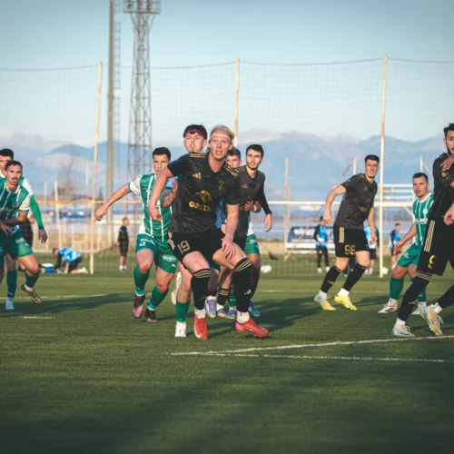 Na zdjęciu widoczna jest scena z meczu piłkarskiego, w której zawodnicy w czarnych i zielonych strojach rywalizują o piłkę na zielonym boisku.