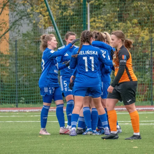 Na zdjęciu widoczna jest grupa kobiet w niebieskich strojach piłkarskich, które celebrują udany moment na boisku, podczas gdy bramkarka w pomarańczowym stroju obserwuje sytuację.