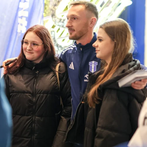 Na zdjęciu dwie dziewczyny pozują z mężczyzną w niebieskim dresie, w tle widać dekoracje świetlne.