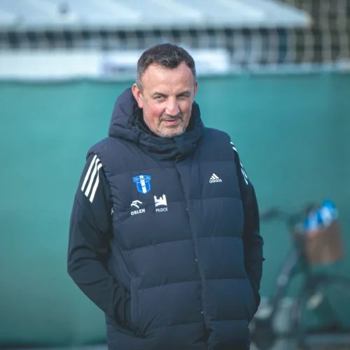 Na zdjęciu widać mężczyznę w ciemnym, ocieplanym bezrękawniku, stojącego na zielonym boisku sportowym.