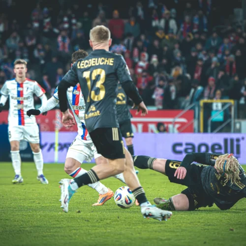 Na zdjęciu widzimy dynamiczną scenę z meczu piłkarskiego, gdzie zawodnik leżący na boisku jest otoczony przez grających kolegów oraz rywali.