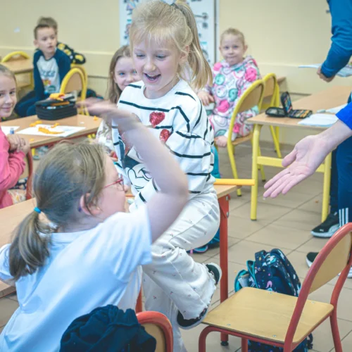 Dzieci w klasie śmieją się i rozmawiają, siedząc przy biurkach w kolorowym otoczeniu.