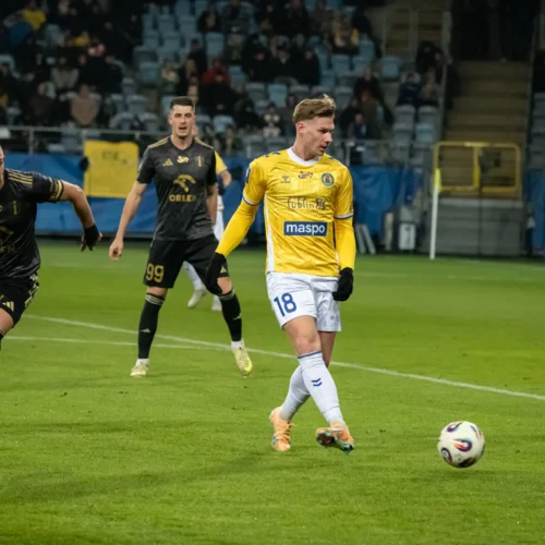 Podczas meczu piłkarskiego zawodnik w żółtej koszulce wykonuje podanie, a dwóch rywali w czarnych strojach obserwuje sytuację.