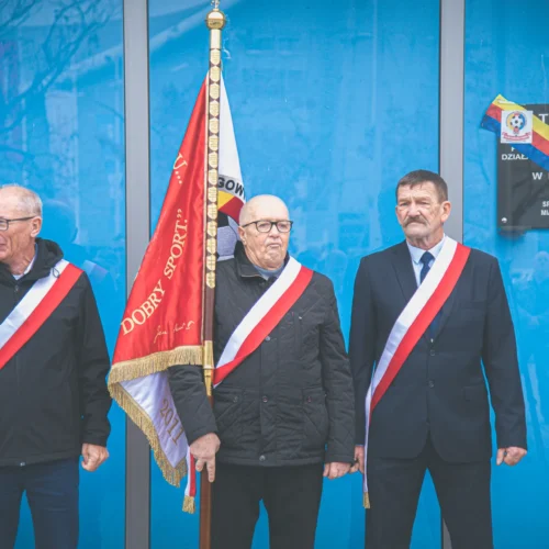 Na zdjęciu trzech mężczyzn w eleganckich ubraniach z wstęgami na ramionach i sztandarem, stojących obok tablicy pamiątkowej w tle.