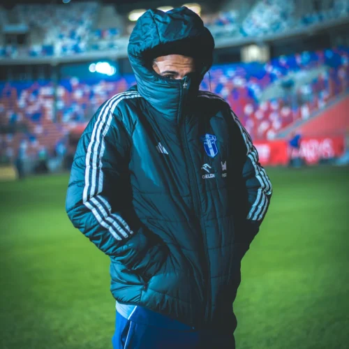 Na zdjęciu widać osobę w dużej, ciemnej kurtce z podniesionym kołnierzem, stojącą na stadionie z piłkarską murawą w tle.