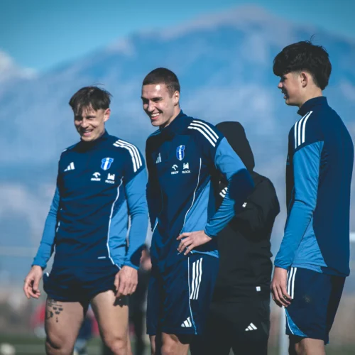 Na zdjęciu trzej młodzi piłkarze w niebieskich strojach sportowych uśmiechają się podczas treningu na boisku, w tle widać górskie krajobrazy.