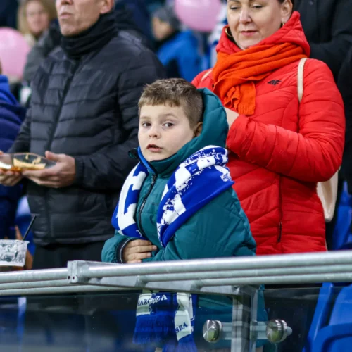 Na zdjęciu widać małego chłopca z kapturem, który stoi na trybunach stadionu, owinięty w szalik, obok niego znajduje się kobieta w czerwonej kurtce.
