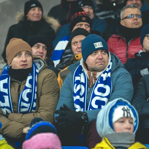 Na zdjęciu widać grupę kibiców na stadionie, którzy oglądają mecz, ubranych w ciepłe kurtki i szaliki w kolorach swojego klubu.