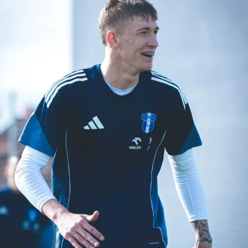 Na zdjęciu widoczny jest młody mężczyzna ubrany w piłkarską koszulkę, który uśmiecha się podczas treningu na świeżym powietrzu.