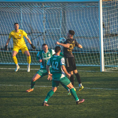 Na zdjęciu widoczna jest intensywna akcja z meczu piłkarskiego, gdzie zawodnik w czarnym stroju strzela na bramkę, a bramkarz w żółtym gotowy jest do obrony.