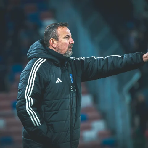 Na zdjęciu widoczny jest trener w czarnej kurtce, który gestykuluje podczas meczu piłkarskiego.