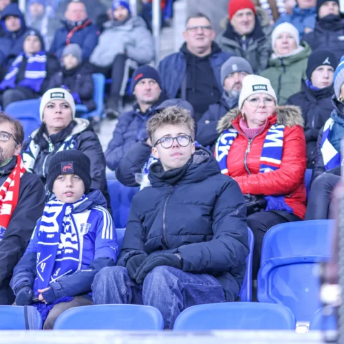Na zdjęciu widoczni są kibice na trybunach, w tym młody chłopiec w niebieskich ubraniach i jego starszy kolega, wszyscy skupieni na wydarzeniu sportowym.