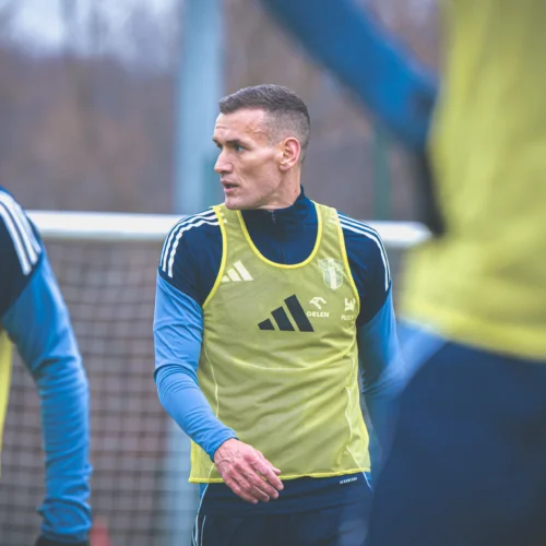 Na zdjęciu widoczny jest mężczyzna w żółtej kamizelce treningowej, skupiony podczas ćwiczeń na boisku, w otoczeniu innych zawodników.