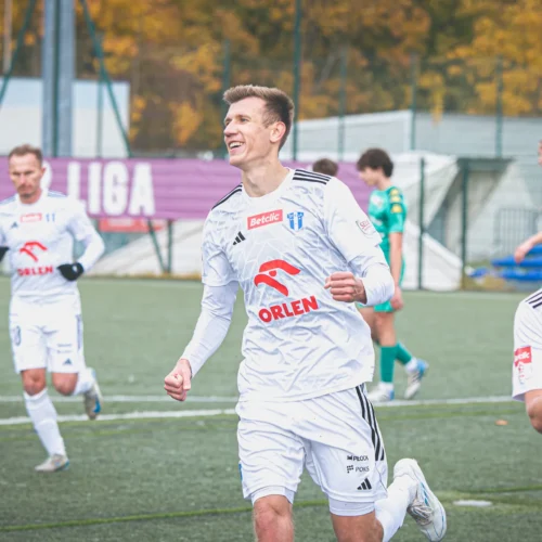 Na zdjęciu widoczny jest radosny piłkarz w białym stroju, biegnący po boisku, podczas gdy w tle znajdują się jego koledzy z drużyny oraz zawodnicy rywalizującej ekipy.