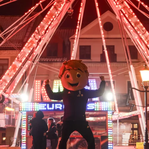 Na zdjęciu widać osobę w dużej, uśmiechniętej masce, stojącą przed diodowym kołem młyńskim ozdobionym kolorowymi światłami, w nocy w radosnej atmosferze.