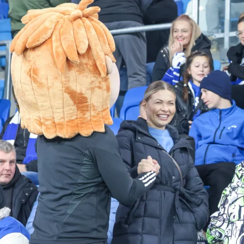 Na zdjęciu widać osobę w kostiumie maskotki oraz uśmiechającą się kobietę wśród kibiców na stadionie.