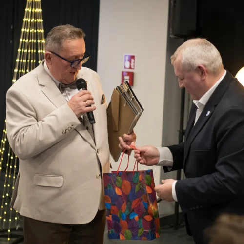 Dwaj mężczyźni w garniturach uczestniczą w wręczeniu prezentu, trzymając mikrofon i kolorową torbę w tle z dekoracyjnym oświetleniem.