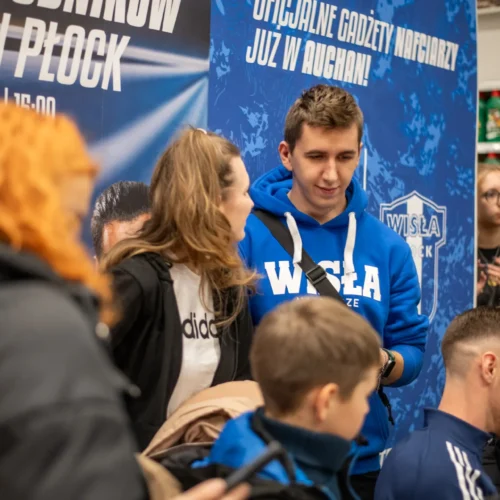Na zdjęciu widać grupę osób w sklepie, z jedną osobą w niebieskiej bluzie z logo Wisły Płock, rozmawiających obok plakatu promującego wydarzenie w Auchan.