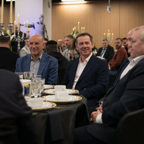 Na zdjęciu widać elegancko ubrane mężczyzn siedzące przy stole podczas uroczystości z dekoracjami i świecami w tle.