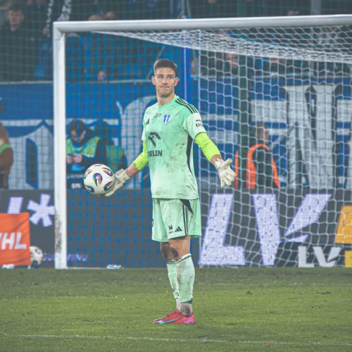 Na zdjęciu widoczny jest bramkarz w zielonym stroju, trzymający piłkę, który stoi na boisku piłkarskim podczas meczu, w tle widać kibiców oraz reklamy.