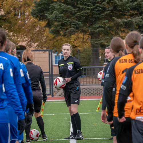 Na zdjęciu widać kobiecą drużynę piłkarską stojącą na boisku, z sędzią trzymającym piłkę wśród zawodniczek w strojach w różnych kolorach.