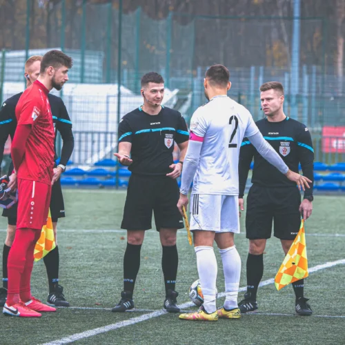 Na zdjęciu widać sędziów oraz kapitanów drużyn rozmawiających przed rozpoczęciem meczu piłki nożnej na boisku.