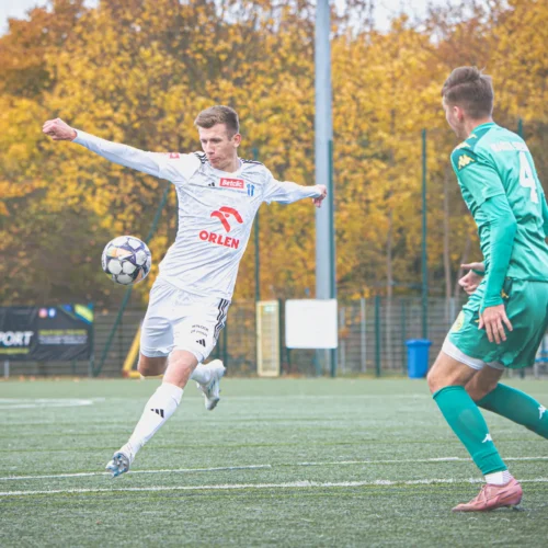 Mężczyzna w białym stroju piłkarskim wykonuje strzał na bramkę podczas meczu, otoczony przez innych zawodników oraz sędziów na boisku.