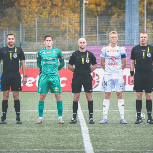 Na zdjęciu widoczni są sędziowie i zawodnicy drużyny piłkarskiej stojący na boisku, gotowi do rozpoczęcia meczu.