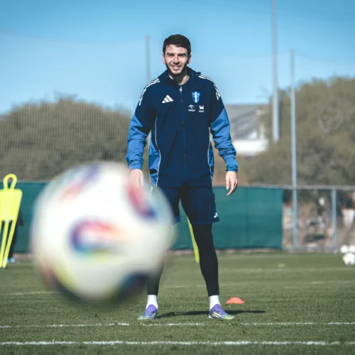 Na zdjęciu widać uśmiechającego się piłkarza w niebieskim stroju sportowym, stojącego na boisku treningowym, w tle widoczna jest bramka oraz kilka piłek.
