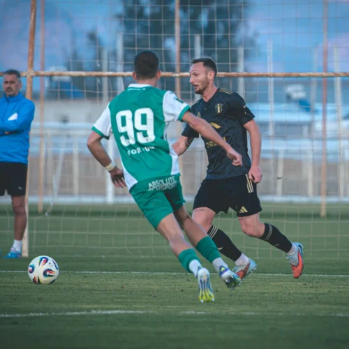 Na zdjęciu dwóch piłkarzy rywalizuje na boisku, podczas gdy jeden z trenerów obserwuje sytuację z boku.