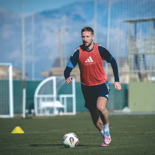 Piłkarz w czerwonym kamizelce treningowej biegnie po boisku, z piłką przy nodze, w tle widać bramki i góry.