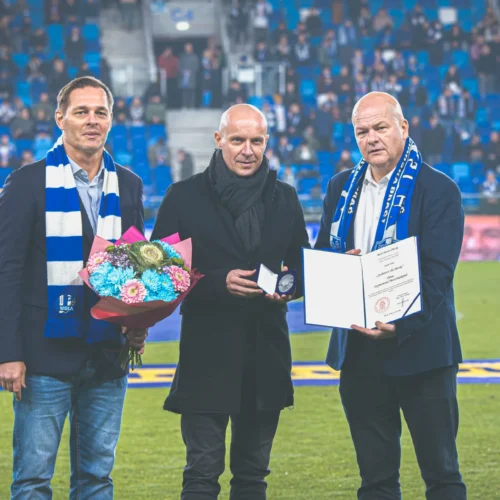 Na zdjęciu trzej mężczyźni stoją na boisku piłkarskim, trzymając kwiaty, medal i dyplom, w otoczeniu kibiców.