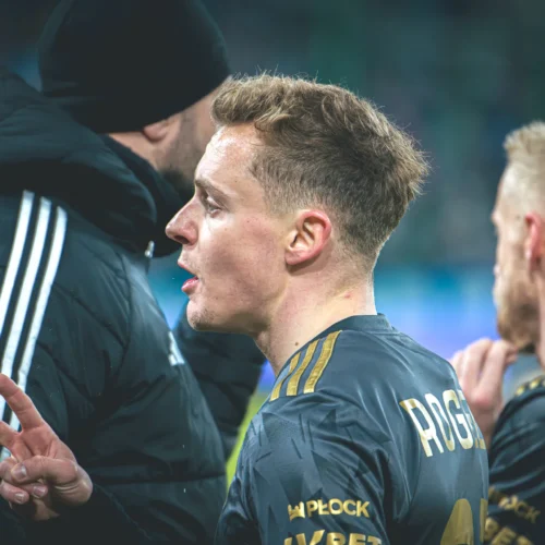 Młody sportowiec w stroju drużynowym gestykuluje, stojąc obok innych zawodników w tle.