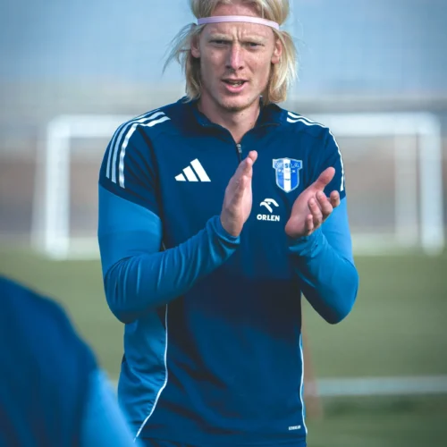 Na zdjęciu widoczny jest mężczyzna w niebieskim stroju sportowym, który klaska i wydaje się być zaangażowany w trening piłkarski na boisku.