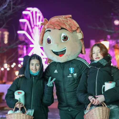 Na zdjęciu widać dwie dziewczyny stojące obok dużej maskotki z uśmiechniętą twarzą, w tle rozświetlone koło młyńskie i ozdoby świetlne.