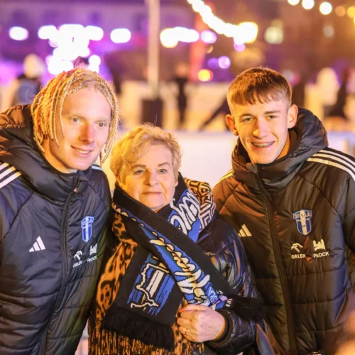 Na zdjęciu dwóch młodych mężczyzn w sportowych kurtkach pozuje z uśmiechniętą kobietą, znajdując się w wieczornym otoczeniu z ozdobnym oświetleniem.