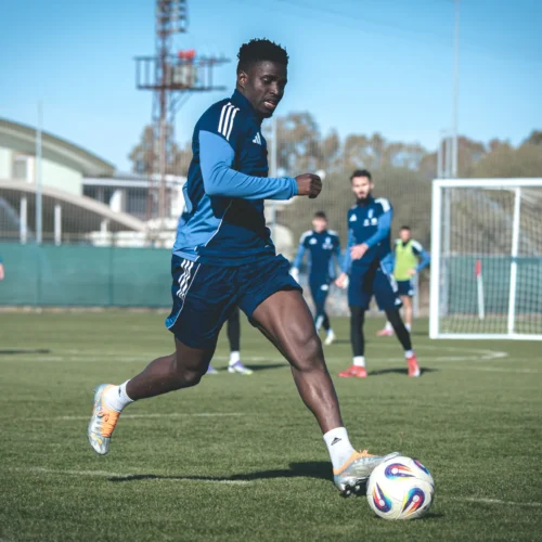 Na zdjęciu widać piłkarza w niebieskim stroju, który biegnie w kierunku piłki podczas treningu na boisku, a w tle znajdują się inni zawodnicy.