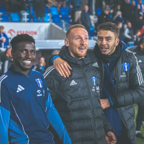 Trzech uśmiechniętych piłkarzy stoi razem na stadionie, w tle widać kibiców.