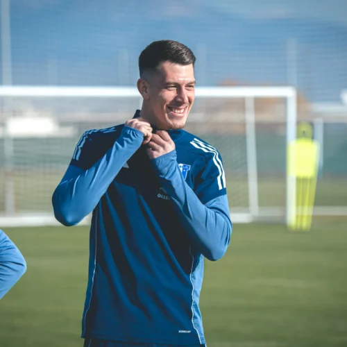 Na zdjęciu widać uśmiechniętego mężczyznę w niebieskim stroju sportowym, który stoi na boisku piłkarskim i poprawia swoje ubranie.